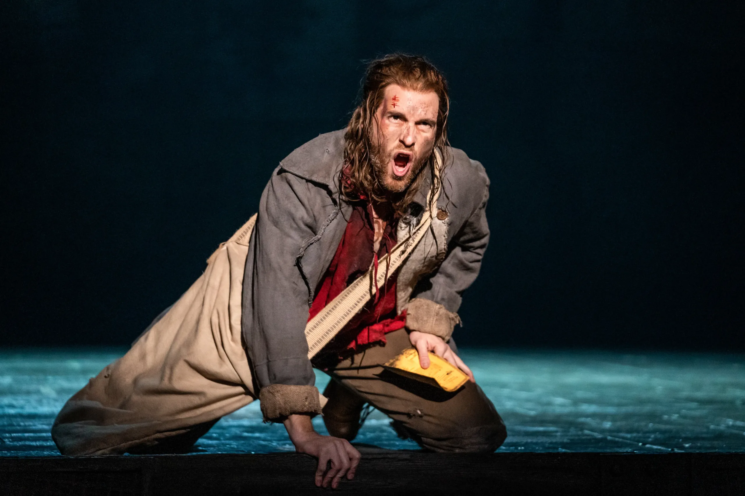 actor with grungy clothing and a scar on his forehead angrily kneeling on the ground