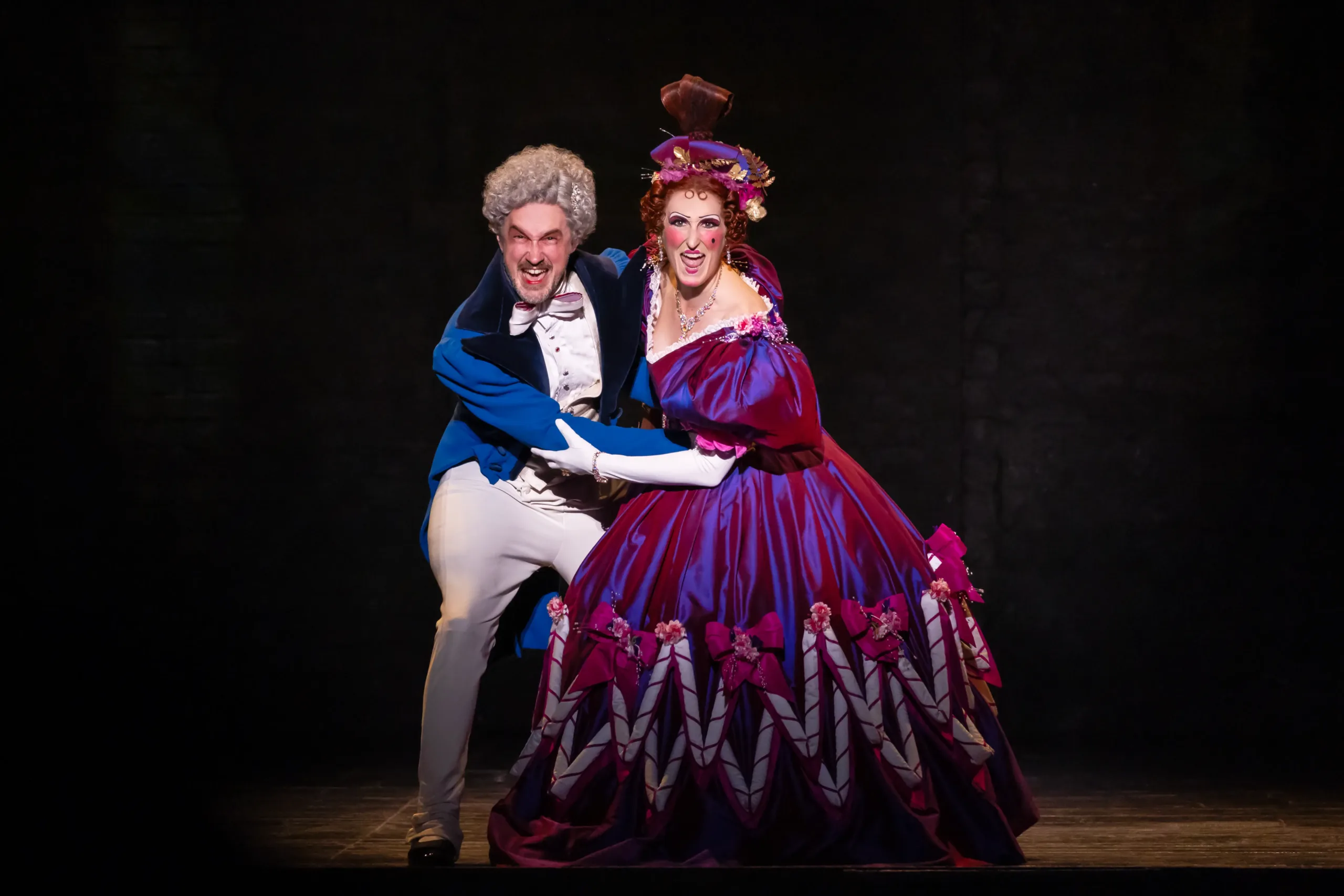 man in a blue suit and woman in a pink dress holding on to each other, looking dramatically at the audience