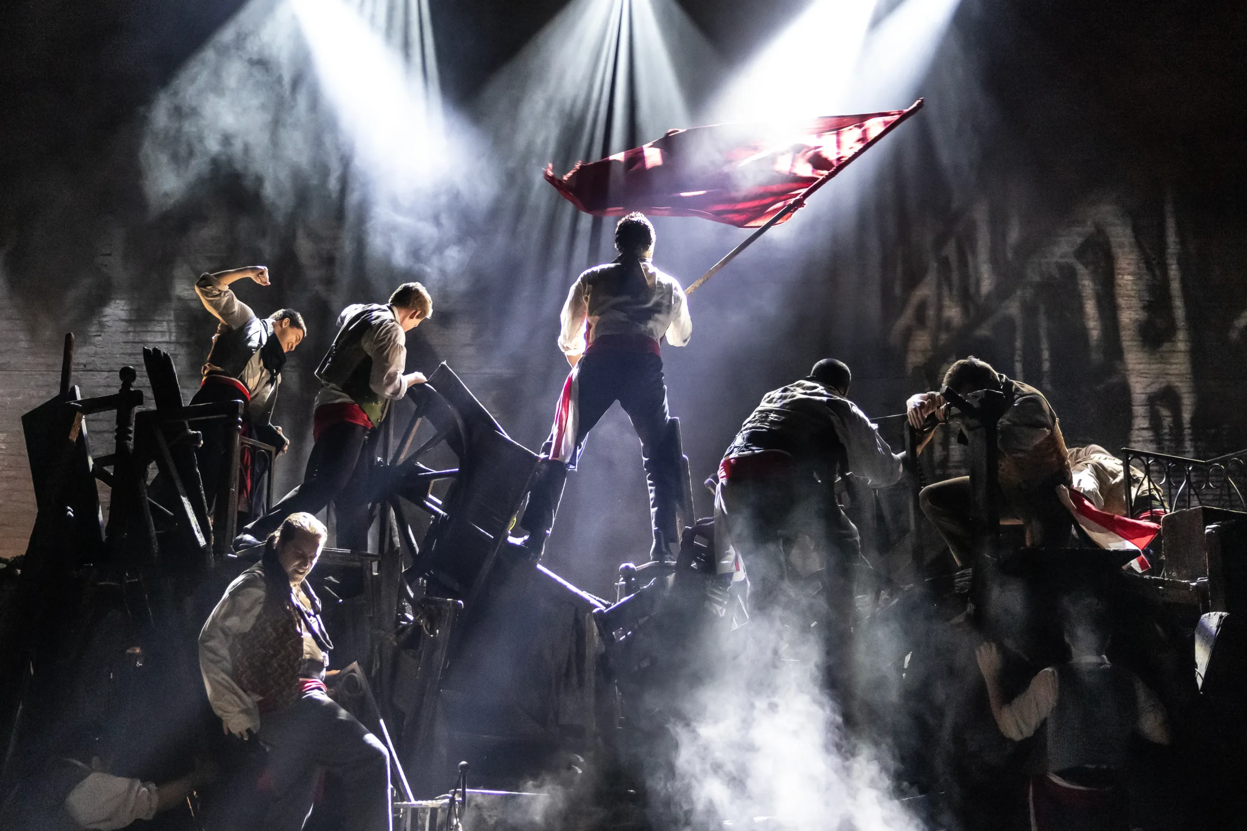 actor surrounded by more actors and a pile of rubble below him, standing triumphantly waving a f