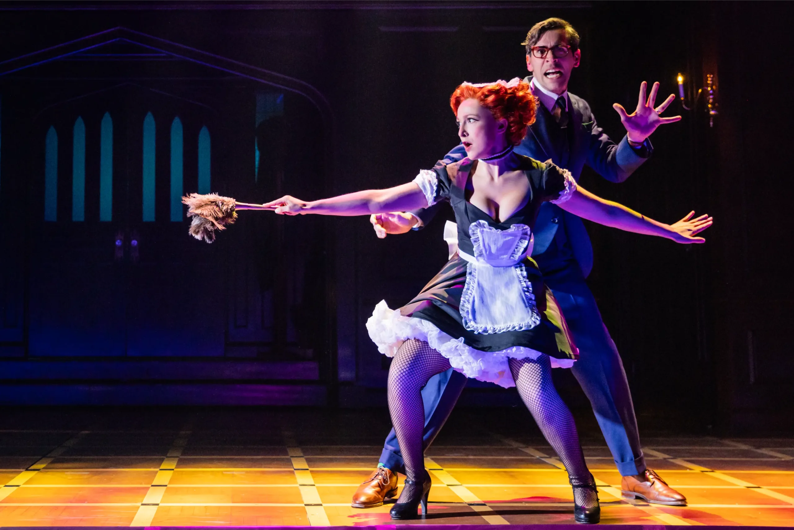 Two actors stand crouched on stage as they are on the lookout for something. A woman in a French maid’s uniform is slowly walking with a feather duster out in front of her as a weapon. A much taller man is behind her looking more frightened than her.