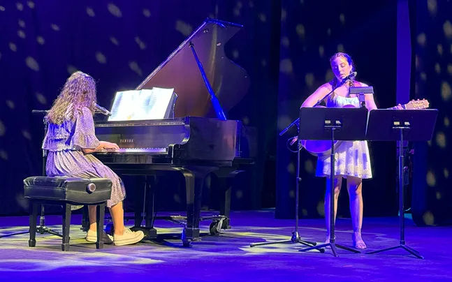 students perform on stage, one a piano and the other playing guitar