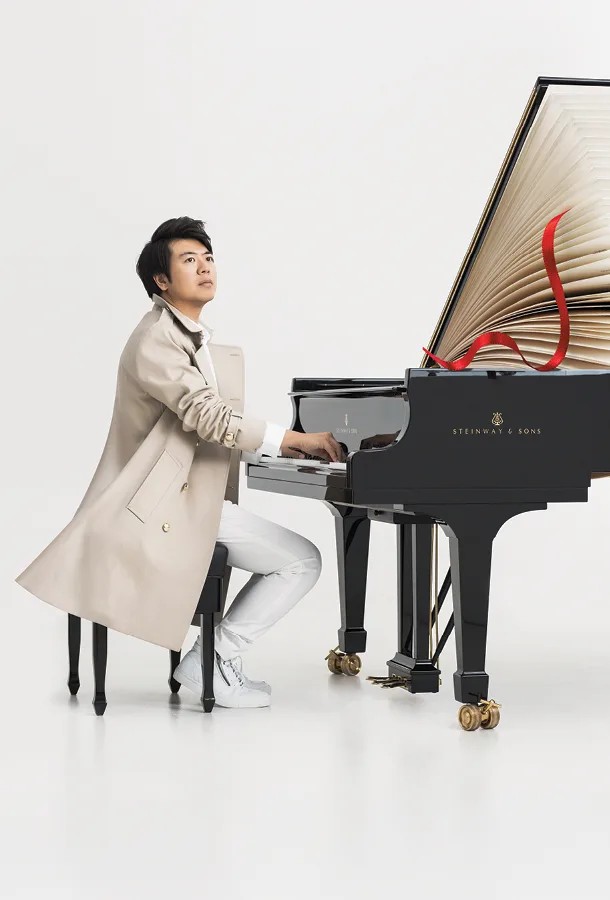 Lang Lang sitting at grand piano wearing light trenchcoat.