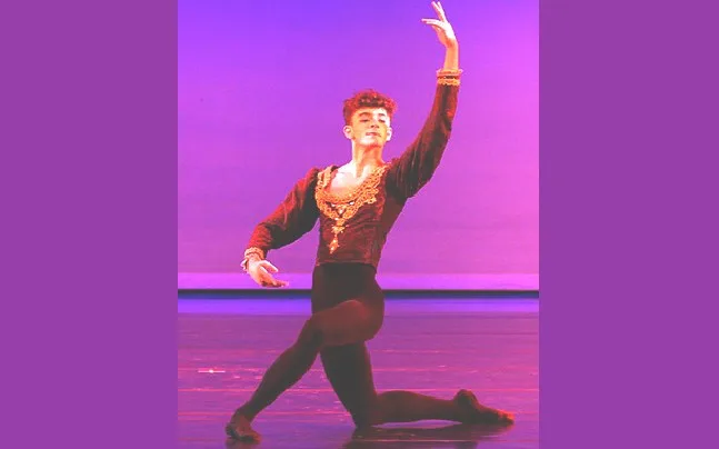 Male dancer kneeling in black costume with arms in fourth position en avant arms