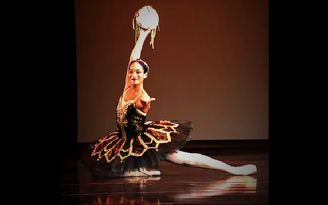 Ballerina kneeling in tutu on stage with leg outstretched and tambourine held above in the air.